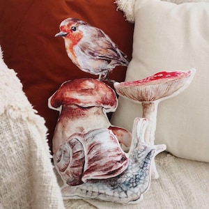Op de afbeelding: Een set van drie decoratieve kussens met een paddenstoel, een slak en een roodborstje. De paddenstoel is rood en wit met een bruine steel. De slak is bruin en wit met een schelp. Het roodborstje is rood, wit en bruin met een gele snavel. De kussens zijn gemaakt van zachte stof en perfect om elke kamer een vleugje whimsy te geven.