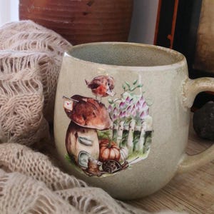 May include: A ceramic mug with a light green glaze features a whimsical illustration of a mushroom house with a bird, a pumpkin, and a snail. The illustration is in shades of brown, orange, red, and green.