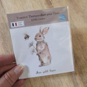 Peut inclure: Une feuille de transfert textile avec une illustration à l'aquarelle d'un lapin brun tenant des marguerites, avec une abeille à proximité. Le texte "Mon petit lapin" est écrit en dessous. L'emballage indique "Transfert Thermocollant pour Tissu".