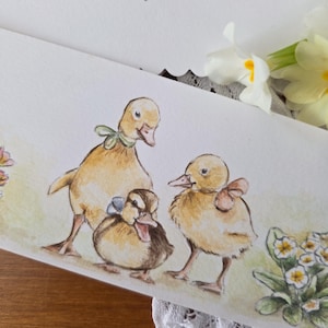 Könnte beinhalten: Aquarellmalerei mit drei Entenküken, jeweils mit einer Schleife. Die gelben und braunen Küken stehen vor einem hellgrünen Hintergrund mit floralen Akzenten. Die Signatur des Künstlers, "Mona Mt", ist sichtbar.