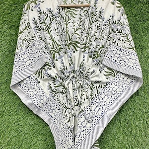 Peut inclure: Kaftan blanc avec un motif floral vert et bleu. Le kaftan est suspendu à un cintre en bois sur un fond vert. Il possède une taille à cordon et une bordure à motifs.