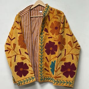 May include: A mustard yellow jacket featuring thick, textured floral embroidery in shades of maroon, orange, and teal. The garment has a stand-up collar, long sleeves, and an interior lining with a vertical white and brown striped pattern.