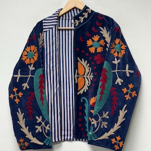 以下が含まれることがあります： 鮮やかなマルチカラーの花と葉の刺繍が施されたネイビーブルーのジャケット。スタンドカラーで、内側は白とネイビーブルーの縦縞模様の裏地になっており、木製のハンガーに掛けられています。