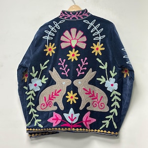 May include: A dark navy blue jacket featuring vibrant, multi-colored embroidery. The design includes two beige rabbits facing each other, surrounded by stylized floral patterns, leaves, and geometric shapes in shades of pink, yellow, and light blue.