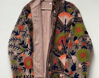 Puede incluir: Una chaqueta de color topo con bordados florales vibrantes en tonos naranja, verde lima y azul marino. La prenda tiene cuello alto, mangas largas y un forro interior de color claro con finas rayas verticales en rosa y marrón.
