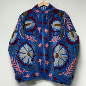 May include: A bright blue velvet jacket featuring intricate floral embroidery in shades of light grey, dark red, brown, and coral pink. The garment has a high collar, long sleeves, and a front opening, showcasing detailed needlework patterns.