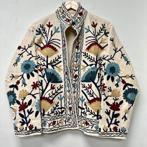 May include: A cream-coloured jacket featuring intricate floral embroidery in shades of blue, red, and yellow. The garment has a high collar, long sleeves, and a front opening, with decorative stitching patterns along the edges and throughout the body.
