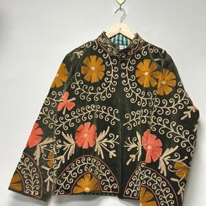 May include: Olive green jacket with floral embroidery in orange and yellow. The jacket features a mandarin collar and is adorned with intricate embroidered patterns. The design includes a mix of floral and scroll motifs, creating a vintage aesthetic.