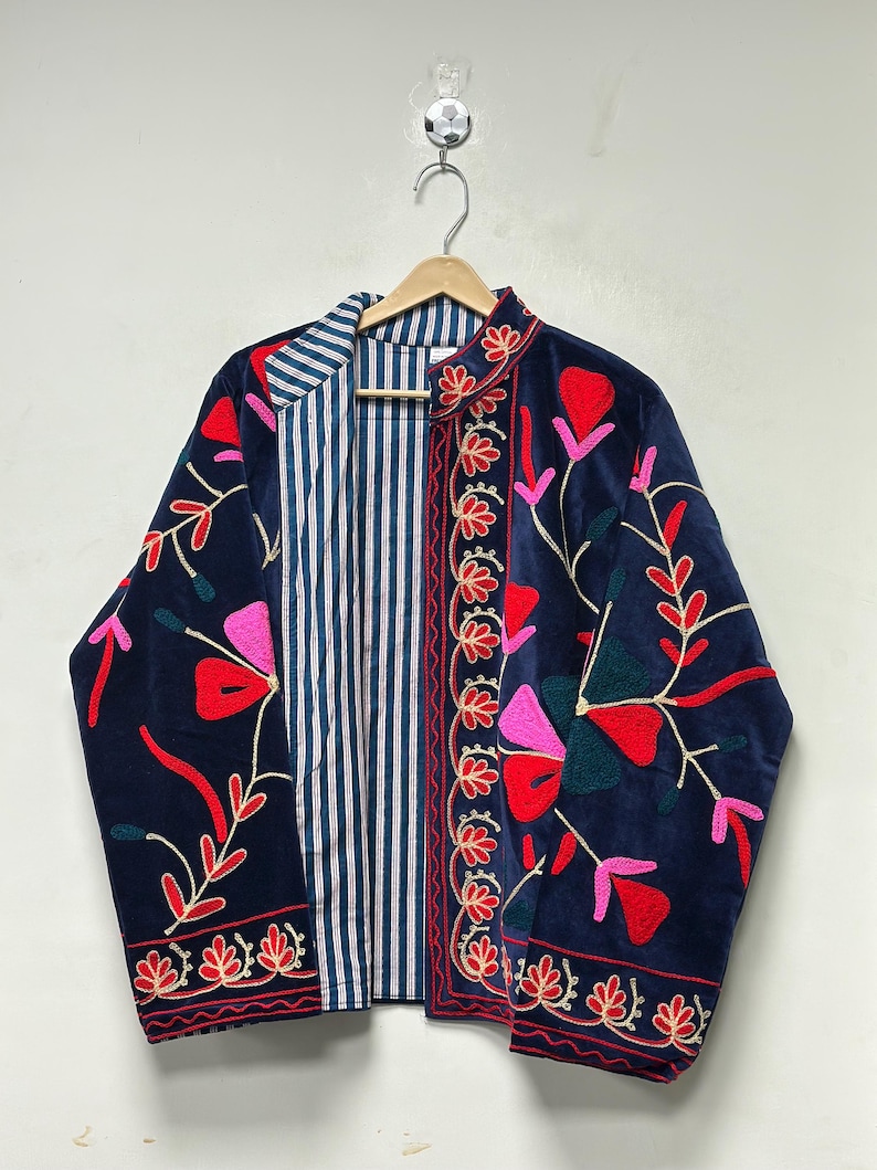 Op de afbeelding: Een marineblauwe jas met levendige bloemenborduursels in rood, roze en donkergroen. De jas heeft een opstaande kraag en een contrasterende binnenvoering met een blauw-wit verticaal strepenpatroon, hangend aan een houten kledinghanger.