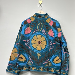 May include: A teal velvet jacket featuring intricate, multi-colored floral and leaf embroidery. The design includes large golden-yellow circular flower motifs, pink petals, and dark brown foliage patterns across the back and sleeves.
