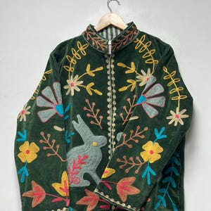 May include: A dark green velvet jacket featuring a front zipper closure and a high collar. The jacket is decorated with colorful embroidered motifs including a light green rabbit, yellow flowers, blue fan-shaped petals, and brown leaves.