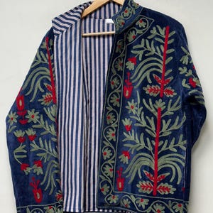 Könnte beinhalten: Eine marineblaue Jacke mit aufwendigen Stickereien in Salbeigrün und Rot. Das Design zeigt florale und botanische Motive auf der Vorderseite und den Ärmeln. Das Innere ist mit einem blau-weiß gestreiften Stoff in vertikalen Linien gefüttert.