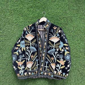 May include: A black jacket with colorful floral embroidery. The jacket features a striped lining and is displayed on a wooden hanger against a green background. The embroidery includes shades of blue, tan, and green.