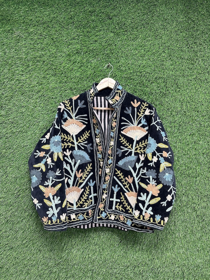 May include: A black jacket with colorful floral embroidery. The jacket features a striped lining and is displayed on a wooden hanger against a green background. The embroidery includes shades of blue, tan, and green.