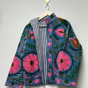 May include: A teal jacket with pink floral embroidery and green leaves. The jacket has a striped lining and a stand-up collar. The jacket is hanging on a wooden hanger.