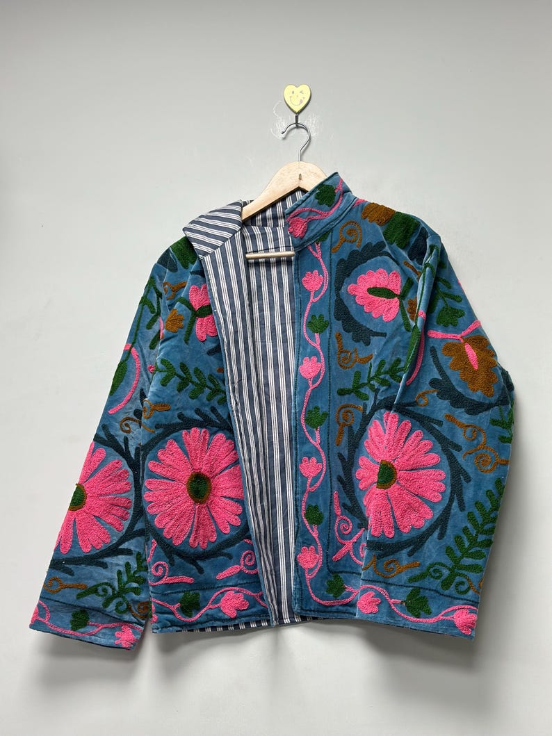 May include: A teal jacket with pink floral embroidery and green leaves. The jacket has a striped lining and a stand-up collar. The jacket is hanging on a wooden hanger.