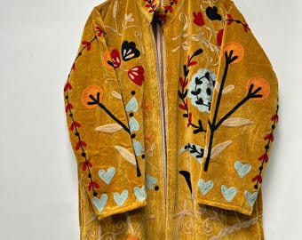Puede incluir: Abrigo largo de terciopelo amarillo mostaza con bordados florales en rojo, negro y azul. El abrigo largo presenta cierre de cremallera y está adornado con corazones y flores bordadas. El abrigo está colgado en una percha de madera.