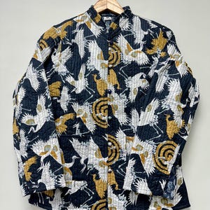 May include: A quilted jacket with a black background and a repeating pattern of white and gold cranes. The jacket has a mandarin collar and button closures. The sleeves are long and the jacket is hanging on a wooden hanger.