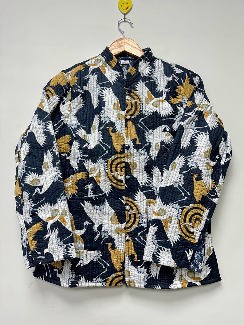 May include: A quilted jacket with a black background and a repeating pattern of white and gold cranes. The jacket has a mandarin collar and button closures. The sleeves are long and the jacket is hanging on a wooden hanger.