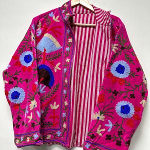 K&ouml;nnte beinhalten: Eine leuchtend pinke Jacke mit aufwendigen Blumenstickereien in Blau-, Rot- und Gr&uuml;nt&ouml;nen. Das Kleidungsst&uuml;ck hat einen hohen Kragen und eine Vorder&ouml;ffnung, die ein Innenfutter mit wei&szlig;en und pinken vertikalen Streifen zeigt.