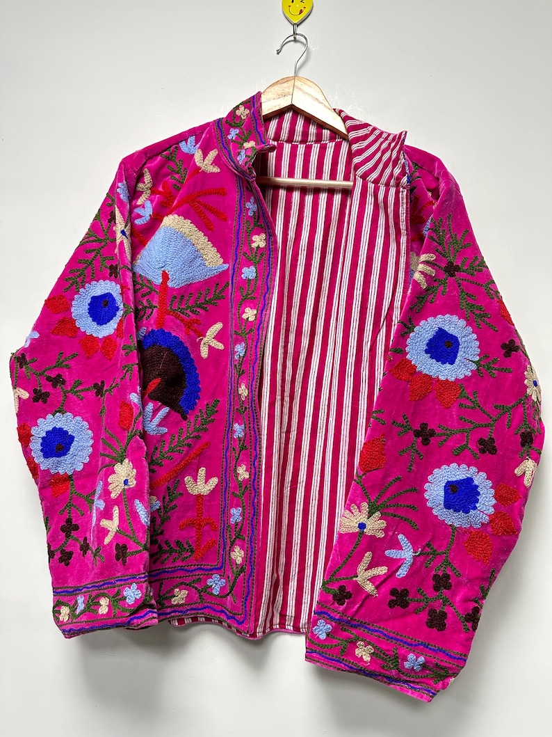 K&ouml;nnte beinhalten: Eine leuchtend pinke Jacke mit aufwendigen Blumenstickereien in Blau-, Rot- und Gr&uuml;nt&ouml;nen. Das Kleidungsst&uuml;ck hat einen hohen Kragen und eine Vorder&ouml;ffnung, die ein Innenfutter mit wei&szlig;en und pinken vertikalen Streifen zeigt.