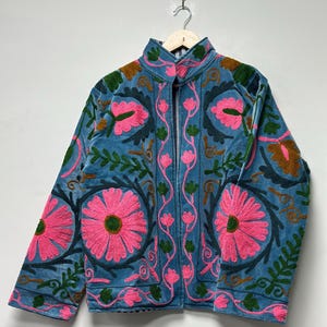 May include: A blue jacket with a high collar and a zipper closure. The jacket is adorned with large pink floral embroidery, green leaves, and brown accents. The jacket is hanging on a wooden hanger.