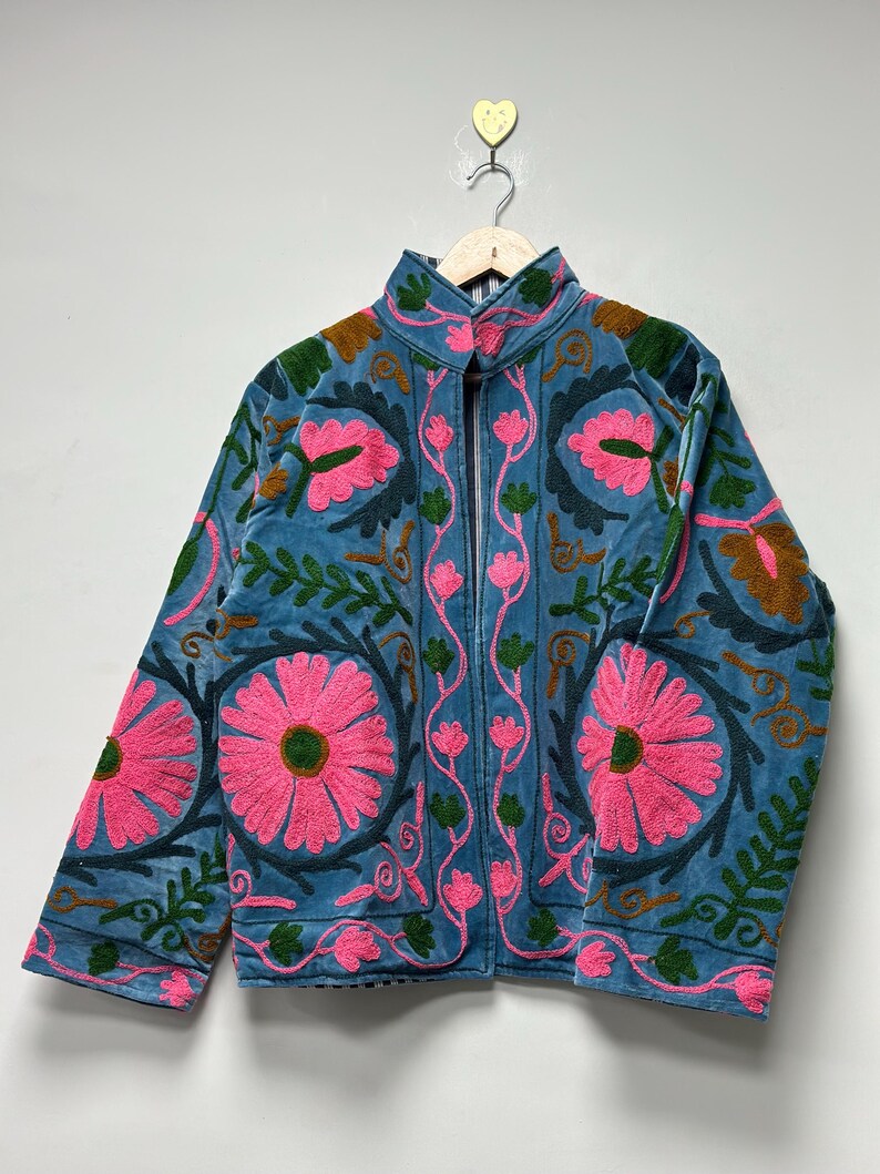 May include: A blue jacket with a high collar and a zipper closure. The jacket is adorned with large pink floral embroidery, green leaves, and brown accents. The jacket is hanging on a wooden hanger.