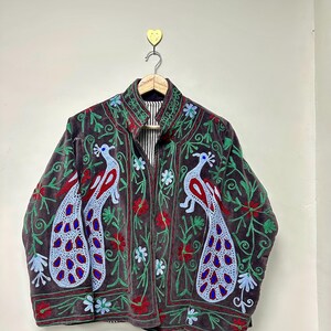 May include: A dark brown velvet jacket with embroidered peacock and floral designs. The jacket features a high collar and a striped lining. The peacock embroidery includes red, blue, and green accents, creating a vibrant and detailed pattern.