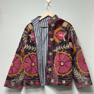 May include: A gray jacket with vibrant floral embroidery in pink, orange, and gold. The jacket features a striped lining and is displayed on a wooden hanger. The embroidery creates a striking contrast against the dark fabric.