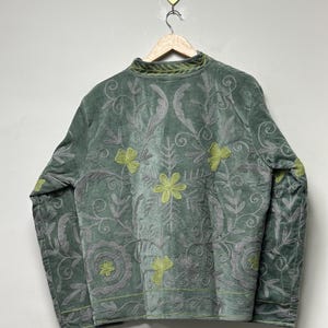 May include: Sage green velvet jacket with intricate floral embroidery in gray and chartreuse. The jacket features a high collar and long sleeves, with detailed patterns across the front and sleeves. The jacket is hanging on a wooden hanger.