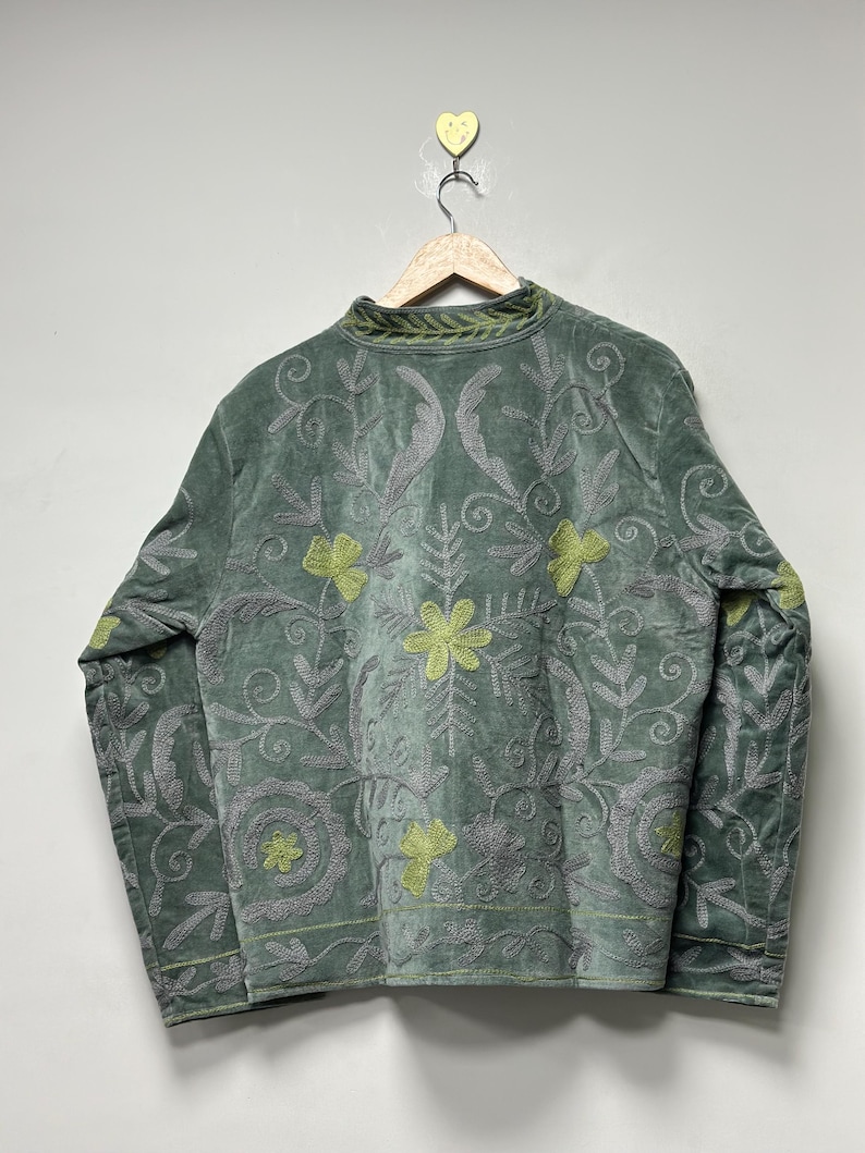 May include: Sage green velvet jacket with intricate floral embroidery in gray and chartreuse. The jacket features a high collar and long sleeves, with detailed patterns across the front and sleeves. The jacket is hanging on a wooden hanger.