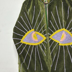 K&ouml;nnte beinhalten: Eine olivgr&uuml;ne Samtjacke mit gestickten Augenmotiven auf den Vorderteilen. Die Augen sind in lavendelfarbenem, pfirsichfarbenem und gelbem Garn gehalten, mit strahlenden Lichtstrahlen in Hellviolett. Der Kragen ist mit gelber Blattstickerei versehen.