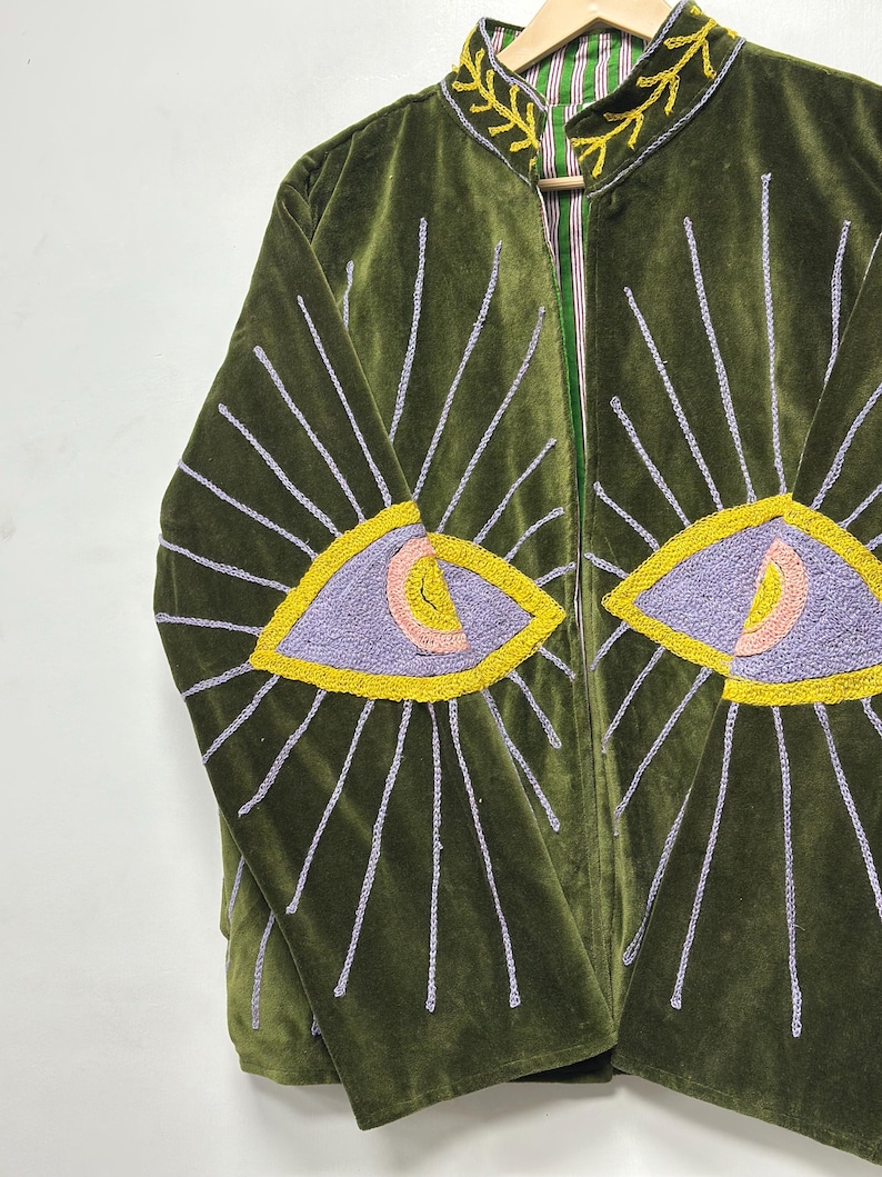 K&ouml;nnte beinhalten: Eine olivgr&uuml;ne Samtjacke mit gestickten Augenmotiven auf den Vorderteilen. Die Augen sind in lavendelfarbenem, pfirsichfarbenem und gelbem Garn gehalten, mit strahlenden Lichtstrahlen in Hellviolett. Der Kragen ist mit gelber Blattstickerei versehen.
