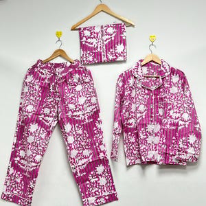 Puede incluir: Un conjunto de pijama de tres piezas con un vibrante estampado floral fucsia y blanco. El conjunto incluye pantalones de pijama, una camisa abotonada de manga larga y una máscara para los ojos a juego. La tela presenta rayas verticales y un diseño floral repetido.