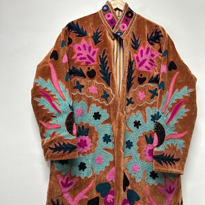 Può includere: Lungo cappotto in velluto marrone con ricami floreali intricati in turchese, rosa e nero. Il cappotto presenta un colletto alla coreana ed è appeso a una gruccia di legno. Il ricamo crea un contrasto sorprendente.