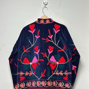 Op de afbeelding: Een donkerblauw fluwelen jasje met levendig bloemenborduursel in tinten rood, roze en donkergroen. Het ontwerp heeft een hoge kraag met decoratieve stiksels en een bijpassende geborduurde rand langs de onderste zoom van het jasje.