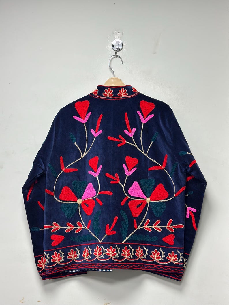 Op de afbeelding: Een donkerblauw fluwelen jasje met levendig bloemenborduursel in tinten rood, roze en donkergroen. Het ontwerp heeft een hoge kraag met decoratieve stiksels en een bijpassende geborduurde rand langs de onderste zoom van het jasje.
