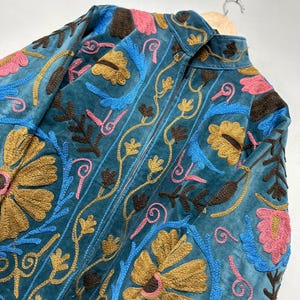 May include: A teal velvet jacket featuring intricate embroidery in shades of mustard yellow, bright blue, dark brown, and pink. The design showcases large floral motifs and scrolling vine patterns across the front, with a high collar and zipper.