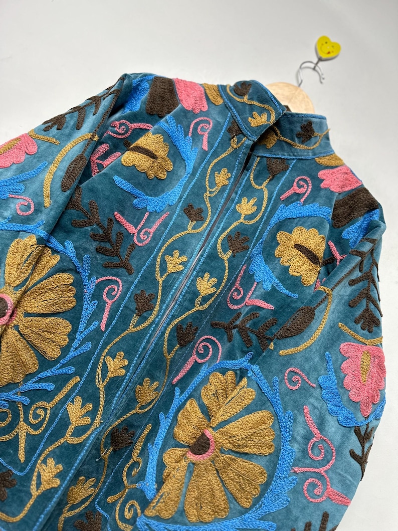 May include: A teal velvet jacket featuring intricate embroidery in shades of mustard yellow, bright blue, dark brown, and pink. The design showcases large floral motifs and scrolling vine patterns across the front, with a high collar and zipper.