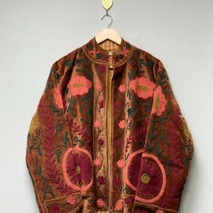 Könnte beinhalten: Braune Samtjacke mit aufwendiger Blumenstickerei in Rot-, Rosa- und Grüntönen. Die Jacke hat einen hohen Kragen, einen durchgehenden Reißverschluss und hängt an einem Holzbügel.