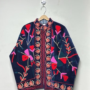 Op de afbeelding: Een marineblauw fluwelen jasje met levendig bloemenborduursel in tinten rood, roze en donkergroen. Het kledingstuk heeft een mandarijnkraag, lange mouwen en een verticale decoratieve rand langs de opening aan de voorzijde met een gestreepte voering.