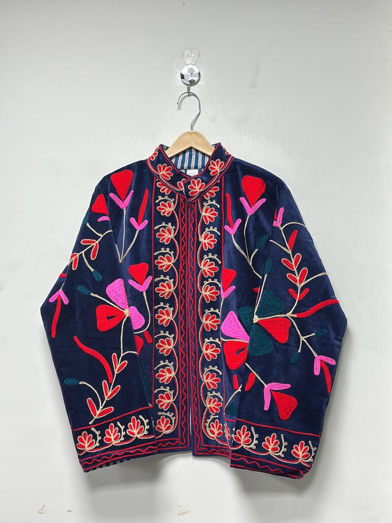 Op de afbeelding: Een marineblauw fluwelen jasje met levendig bloemenborduursel in tinten rood, roze en donkergroen. Het kledingstuk heeft een mandarijnkraag, lange mouwen en een verticale decoratieve rand langs de opening aan de voorzijde met een gestreepte voering.
