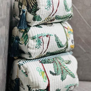May include: Three white quilted makeup bags with a green and brown tropical print. The bags are stacked on top of each other. Each bag has a blue tassel.