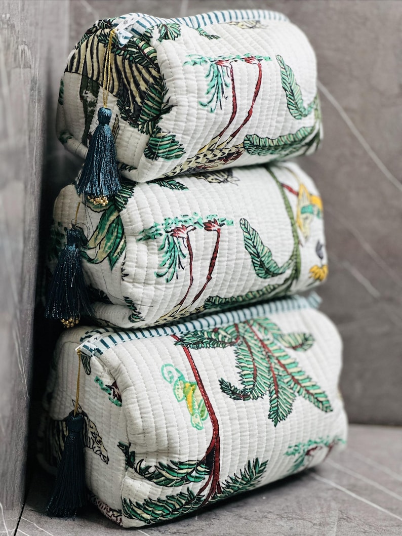 May include: Three white quilted makeup bags with a green and brown tropical print. The bags are stacked on top of each other. Each bag has a blue tassel.