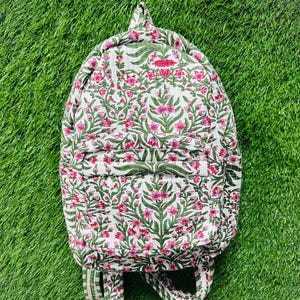 May include: A floral-patterned backpack with a white background and pink and green floral design. The backpack has a top handle, adjustable straps, and a front pocket. The backpack is made of a quilted fabric.