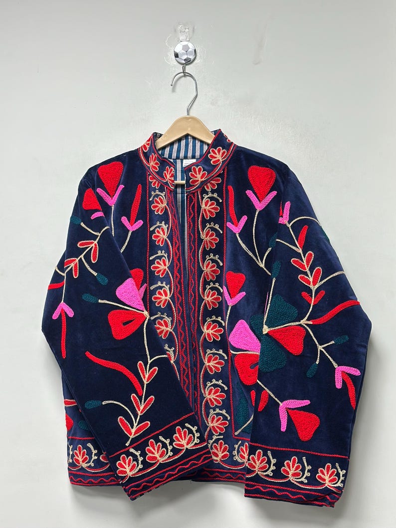 Op de afbeelding: Een marineblauw fluwelen jasje met levendig bloemenborduursel in tinten rood, roze en donkergroen. Het kledingstuk heeft een opstaande kraag, lange mouwen en een opening aan de voorzijde met verticale geborduurde patronen langs de sluiting.