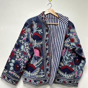 Könnte beinhalten: Blaue Samtjacke mit aufwendiger Blumenstickerei in Rot, Rosa und Türkis. Die Jacke hat ein gestreiftes Innenfutter in Marineblau und Weiß. Die Jacke hängt an einem Holzkleiderbügel.