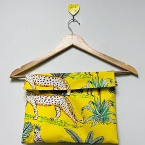 Puede incluir: Bolso de mano amarillo con cierre de solapa, con un estampado repetido de leopardos y plantas tropicales. La bolsa est&aacute; colgada de una percha de madera. El estampado de leopardo tiene manchas blancas sobre fondo beige.