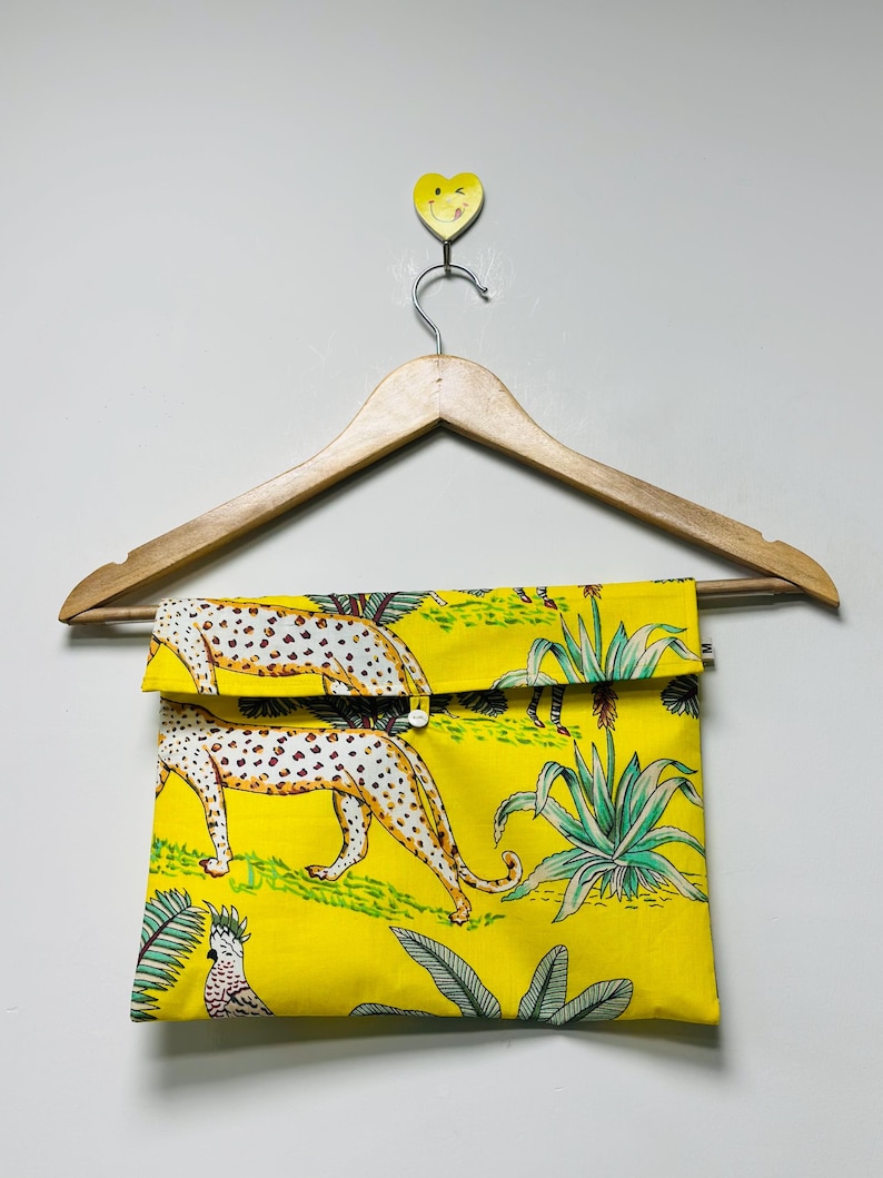 Puede incluir: Bolso de mano amarillo con cierre de solapa, con un estampado repetido de leopardos y plantas tropicales. La bolsa est&aacute; colgada de una percha de madera. El estampado de leopardo tiene manchas blancas sobre fondo beige.