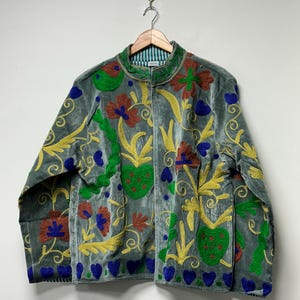 May include: A grey velvet jacket featuring vibrant, multi-colored embroidery in shades of yellow, green, blue, and rust. The design includes floral and heart-shaped motifs across the front and sleeves, with a high collar and a striped inner lining.